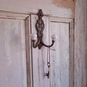 Add vintage old-world charm to any room with our Antiqued Brass Double Hook with Porcelain Knobs. This solid cast iron piece features and aged brass finish and an ornate floral design. Includes two hooks capped with vintage porcelain knobs.