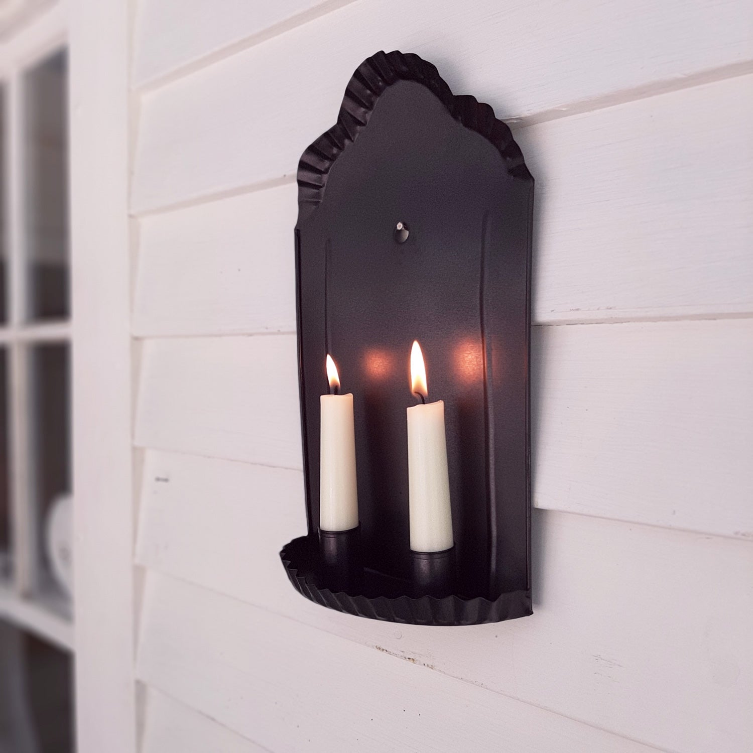 Our Carriage House Candle Wall Sconce is a black metal taper holder featuring decorative fluted edges around the rim of the pan and the top of the piece.. This black metal wall sconce holds two taper candles. Its simple design works well with rustic farmhouse or  coastal cottage decor.  7”L x 2.25”W x 12”H