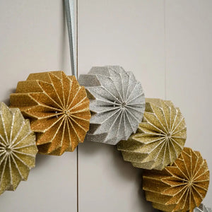 This handmade Sparkle Paper Pinwheel Wreath is an eye-catching and unique holiday accent. The chunky paper pinwheel style flowers sparkle with a combination of glittery gold, silver and copper. The wreath is topped off with a silvery-grey ribbon for hanging. This charming wreath lends a down-to-earth, handmade feel to your holiday home. Made with heavy-duty cardboard. For indoor use only.  15" diam