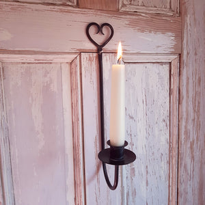 The Rustic Heart Taper Candle Holder adds primitive farmhouse charm to any room. The relaxed design features a metal taper holder with an aged, black/gray finish. The top of the holder features a beautiful heart shape and a pre-drilled hole for hanging. The holder curves up at the base and holds a candle tray with a taper candle cup. Measures 12" high by 2" wide with a ⅞" candle cup.